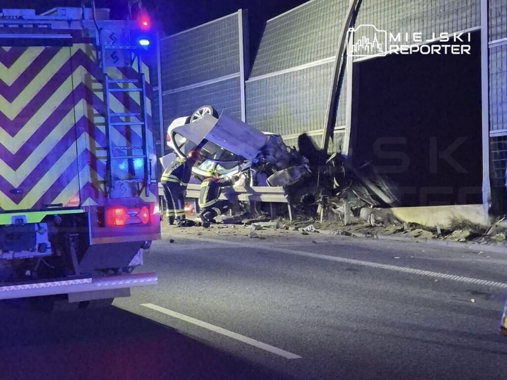 Strażacy pracują nad wywróconym samochodem osobowym przy drodze, obok ogrodzenia z paneli słonecznych.