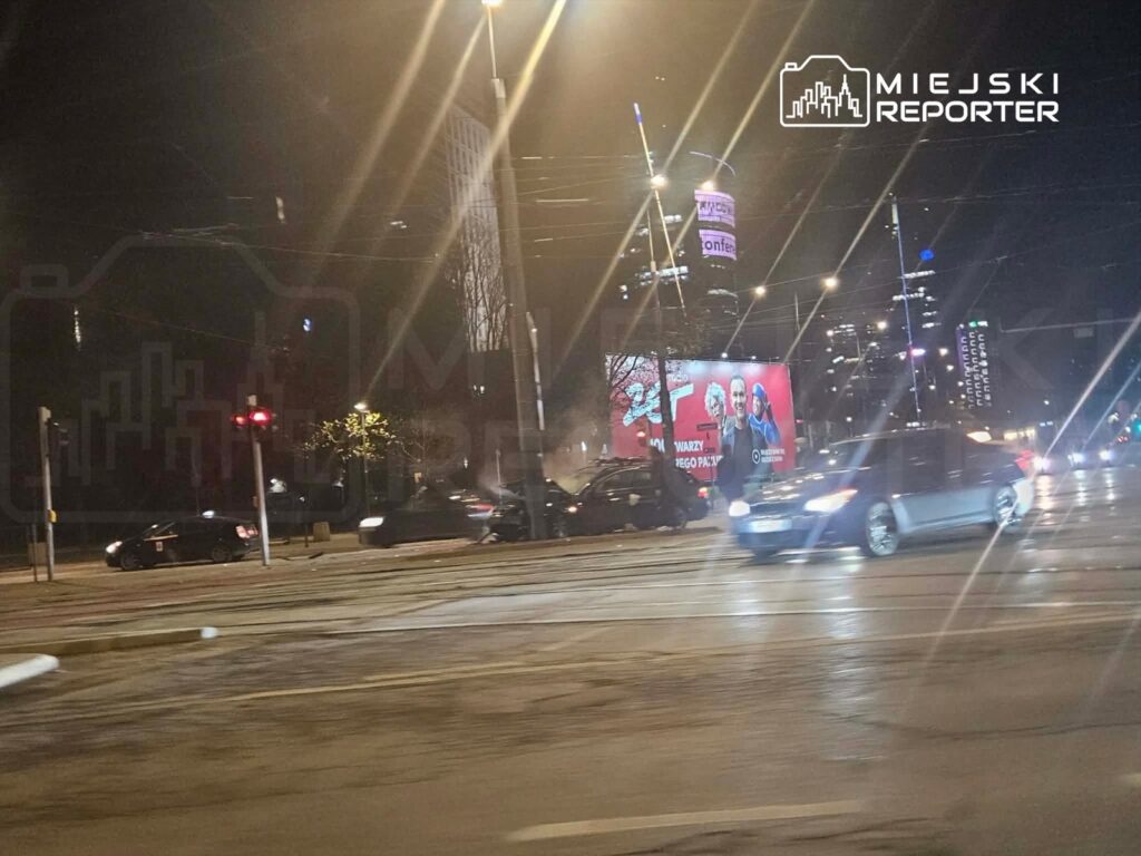 Ruch uliczny w nocy, z samochodami zatrzymującymi się na czerwonym świetle, w tle widoczny billboard reklamowy.