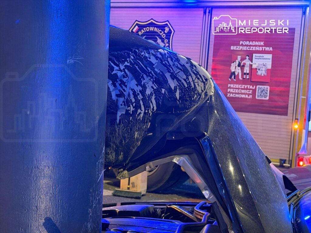 Uszkodzone auto osobowe uderzyło w metalowy słup, zniszczona karoseria widoczna z bliska w nocy. W tle widoczny pojazd ratunkowy.