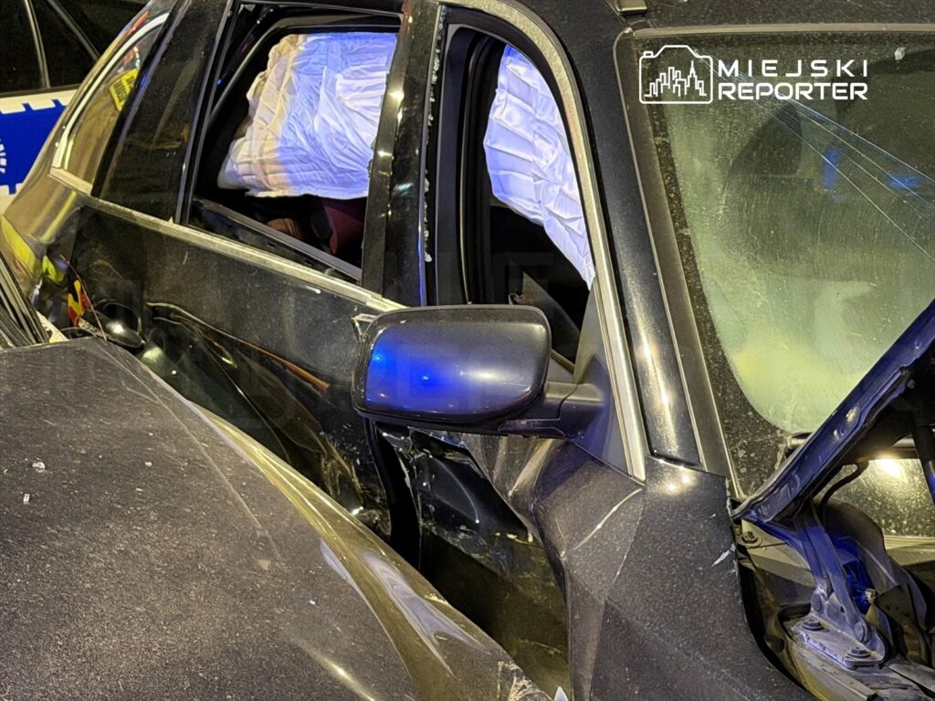 Uszkodzony samochód osobowy z wybitymi szybami, w którym widać poduszkę powietrzną, zaparkowany w nocy.