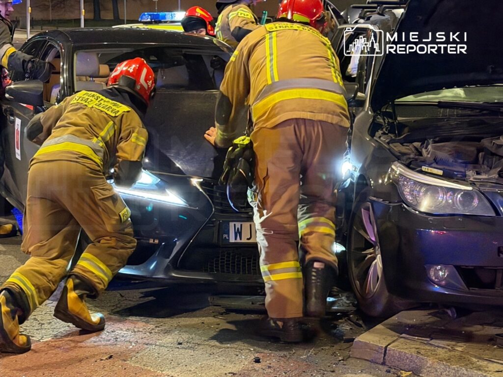 Grupa strażaków w żółtych mundurach pracuje przy rozbitym samochodzie osobowym na ulicy, oświetlonym przez latarnie.
