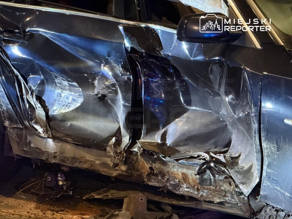 Uszkodzony samochód osobowy z wgniecioną karoserią, stojący na poboczu drogi w nocy, oświetlony przez policyjne światła.