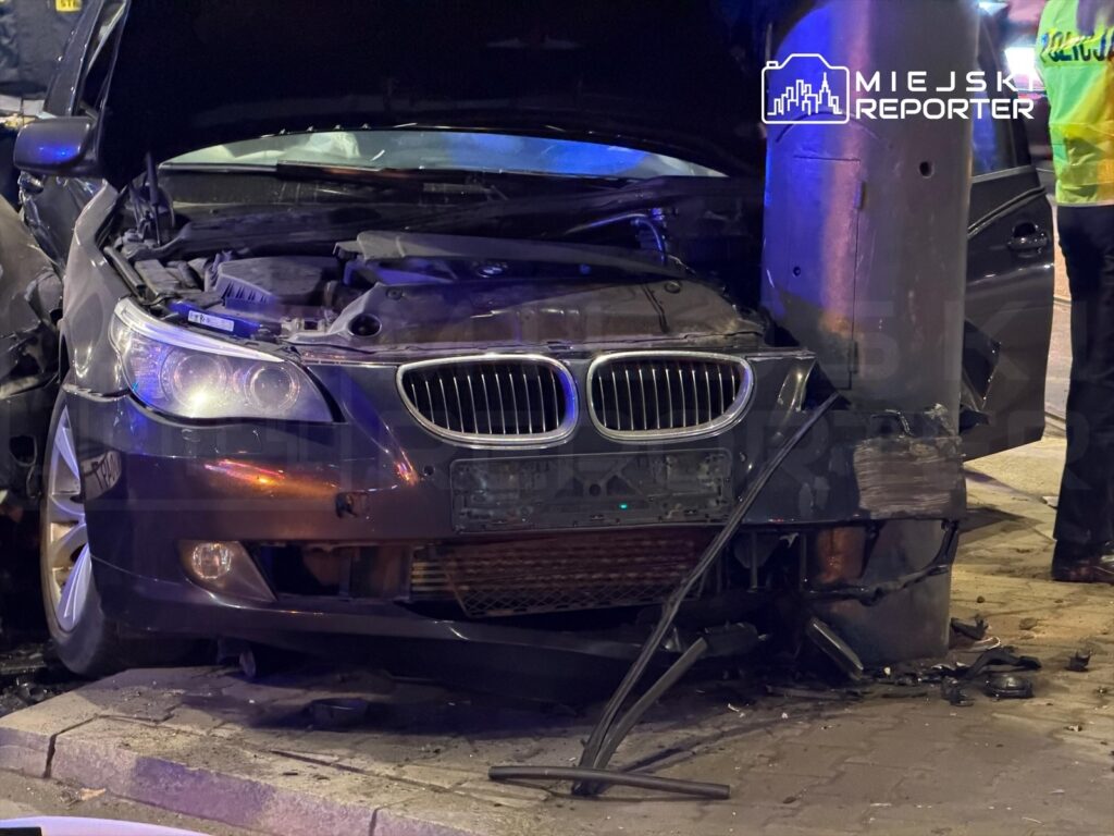 Czarny samochód osobowy uderzył w betonowy słup, zniszczona maska i uszkodzona przednia część pojazdu.