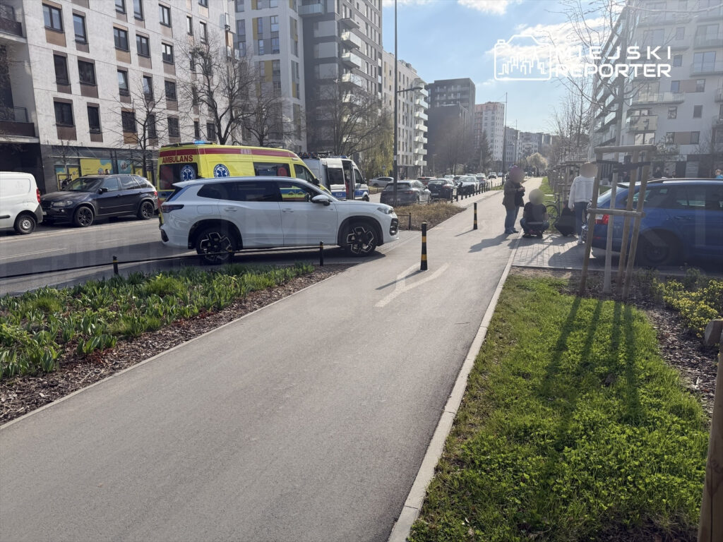 Na chodniku obok drogi stoi biała limuzyna, a obok niej grupka osób rozmawia. W tle widać karetki i nowoczesne budynki mieszkalne.