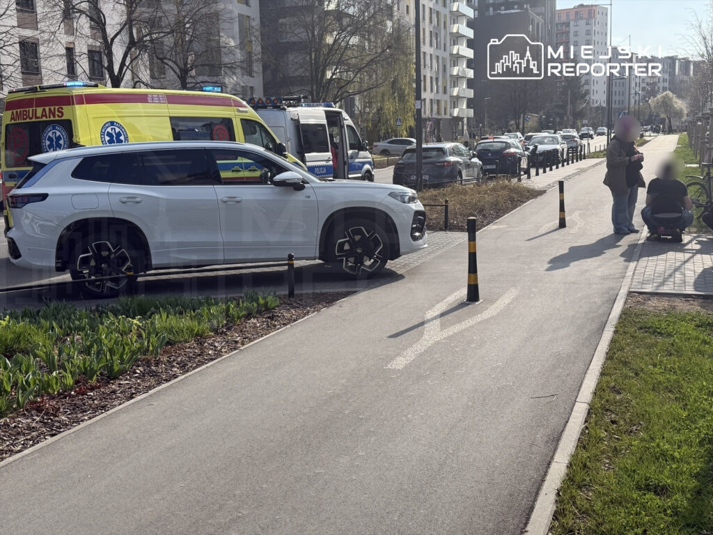 Biały SUV stoi obok karetki, a dwie osoby rozmawiają na chodniku w miejskim otoczeniu.