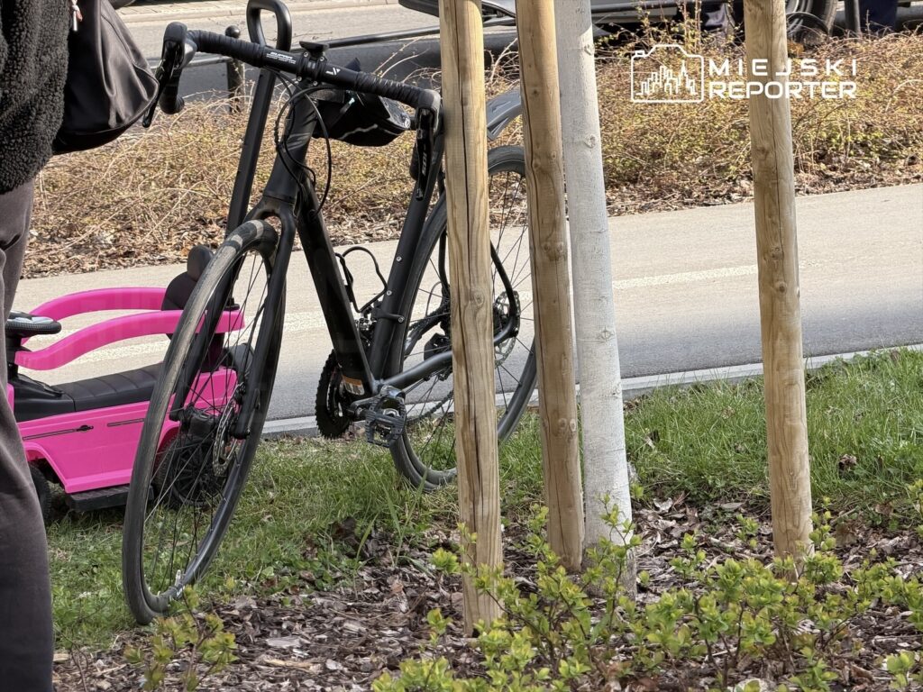 Rower stoi oparty o drewnowy słup w pobliżu trawnika, obok widoczny różowy wózek dziecięcy.