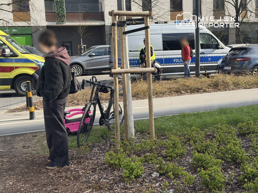 Kobieta w czarnej kurtce stoi obok roweru, w tle widoczny radiowóz policyjny i karetka.