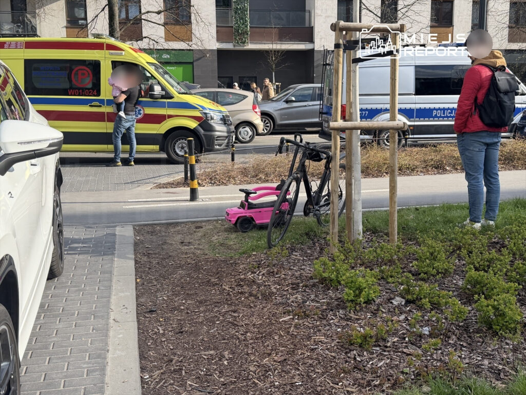 Mężczyzna trzymający dziecko stoi obok karetki pogotowia, a w tle widoczni są przechodnie i radiowóz policji.