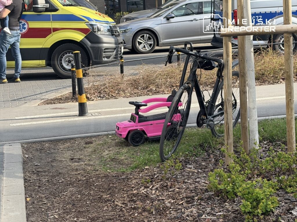 Mężczyzna trzymający dziecko stoi obok roweru, obok znajduje się różowy pojazd dla dzieci na trawniku.