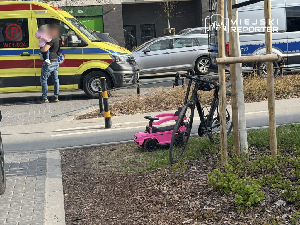 Mężczyzna trzymający dziecko rozmawia z ratownikami przy karetce, obok stoi czarna rower i różowy wózek dla dzieci.