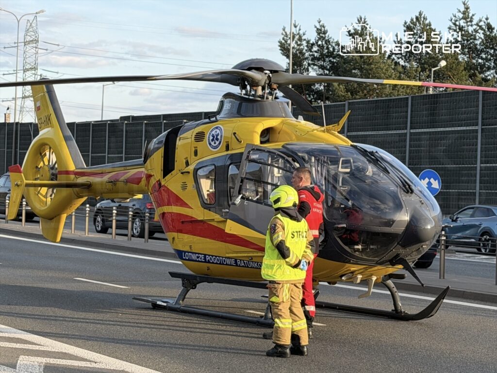 Ratownicy medyczni przygotowują się do akcji obok helikoptera Lotniczego Pogotowia Ratunkowego na ruchliwej drodze.