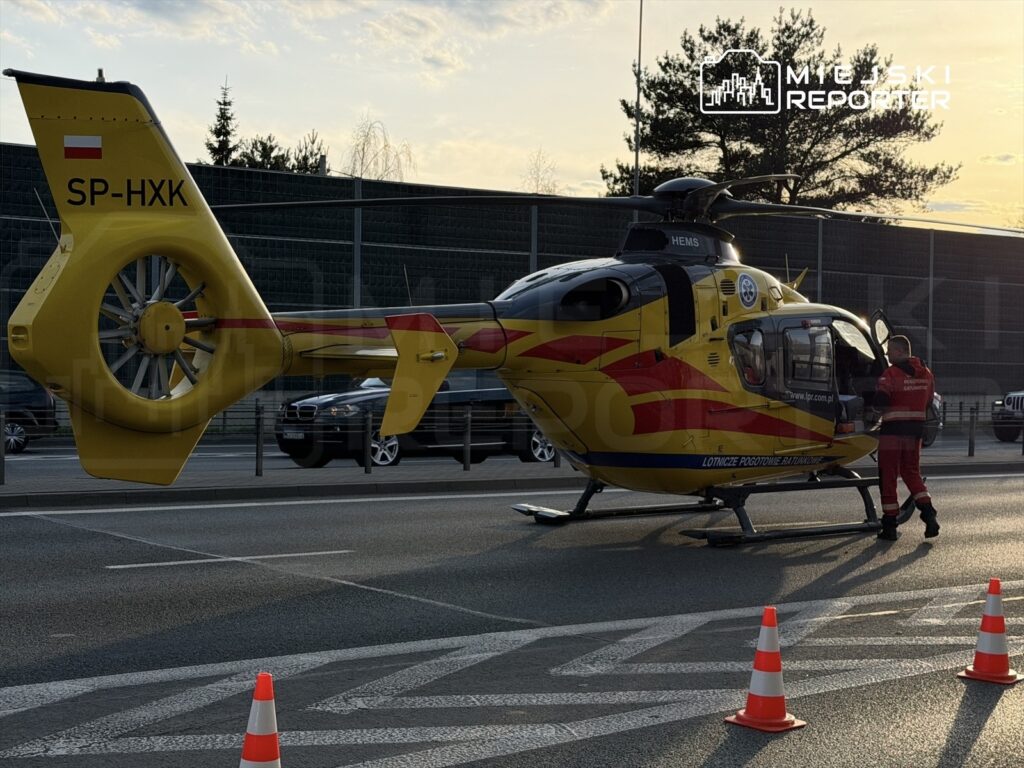 Żółty helikopter ratunkowy lądował na jezdni, obok stoi ratownik w czerwonym stroju, a w tle widać samochody.