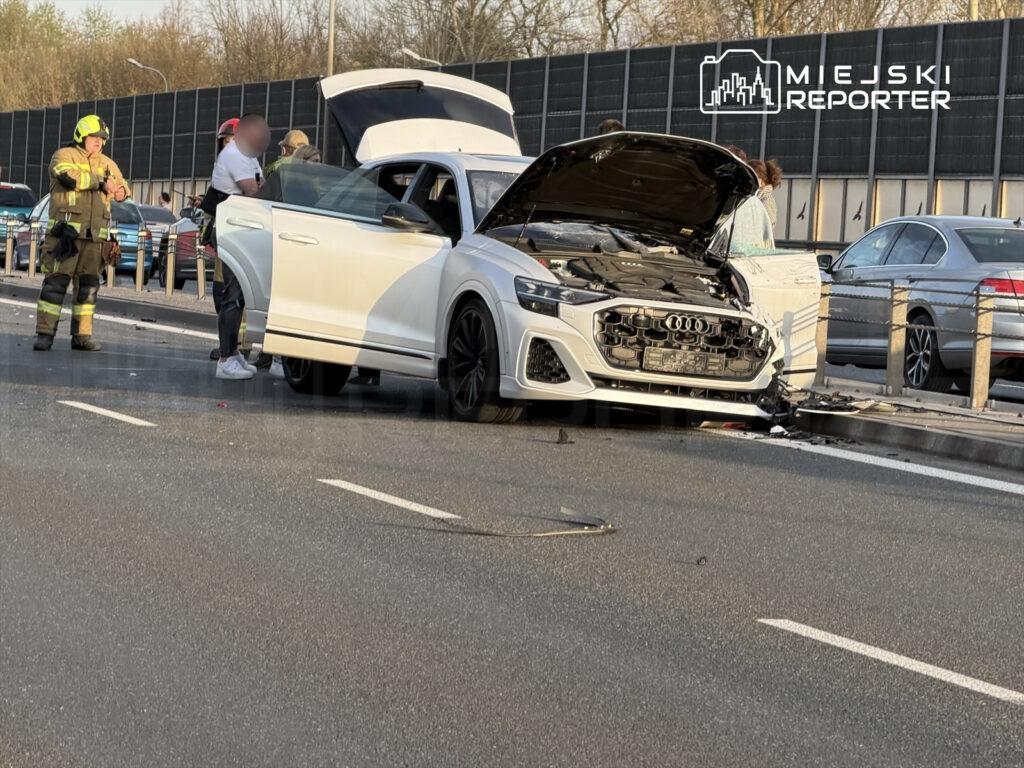 Biały samochód osobowy z uszkodzoną przednią częścią stoi na drodze, obok grupy ratowników w mundurach.