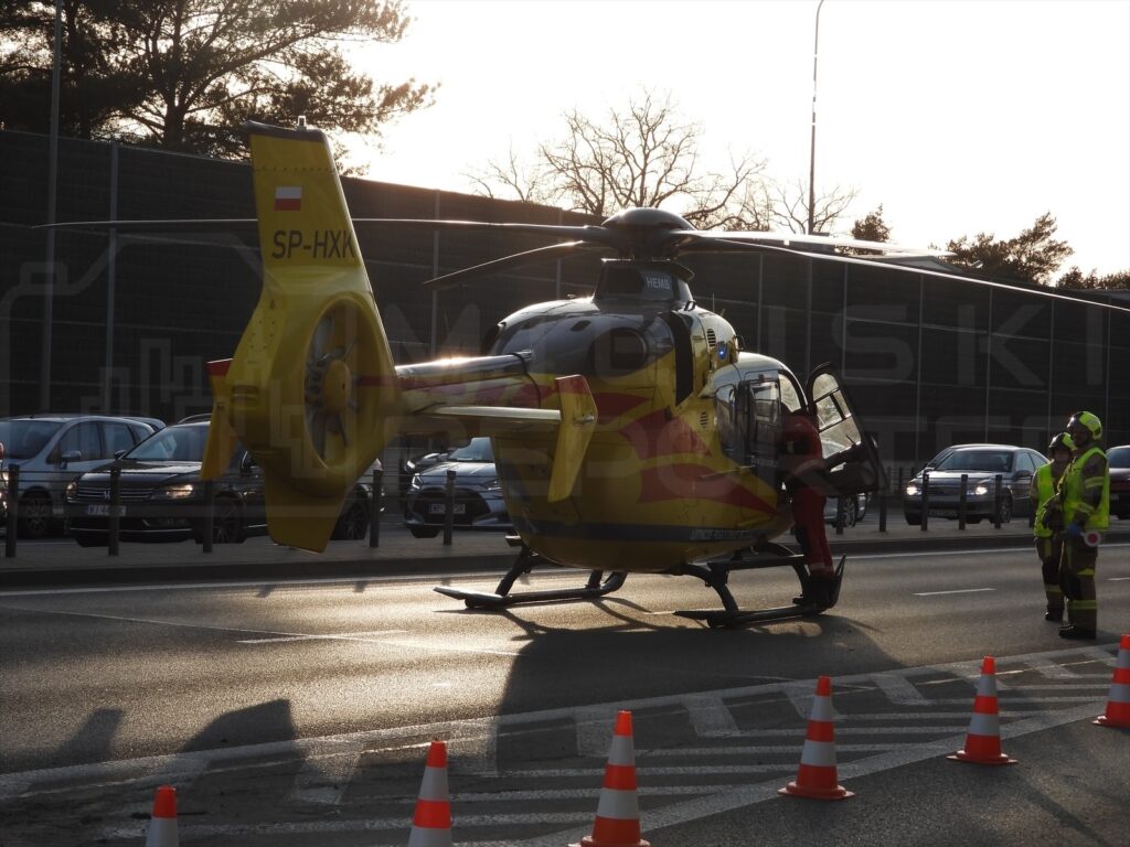 Śmigłowiec ratunkowy ląduje na jezdni, obok grupy ratowników w odblaskowych ubraniach, z ustawionymi pachołkami.