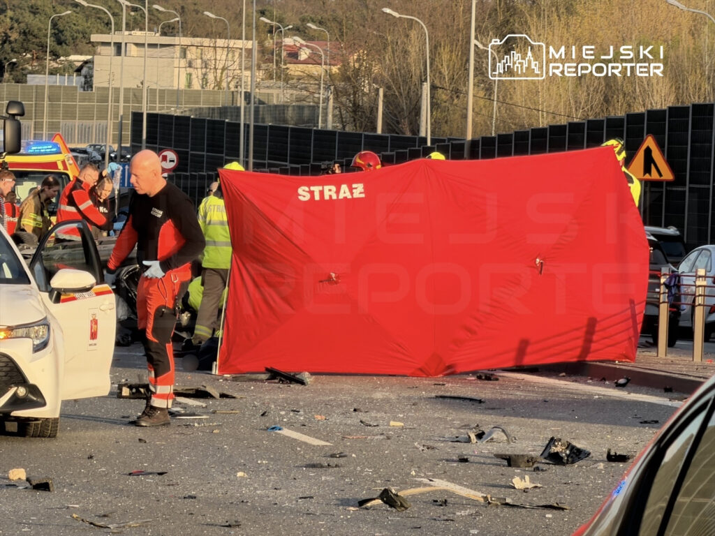 Ratownicy medyczni pracują na miejscu wypadku drogowym, otoczeni przez pojazdy i zniszczone elementy.