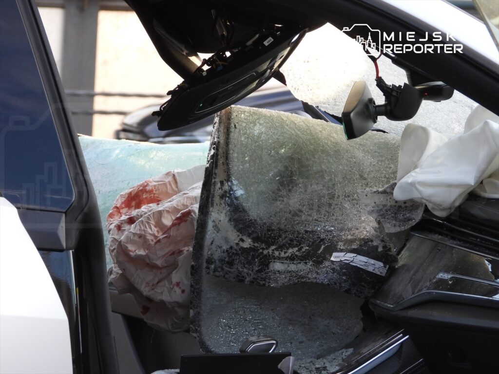 Zniszczony samochód z rozbitymi szybami, wewnątrz widoczny krwawy materiał oraz poduszka powietrzna.