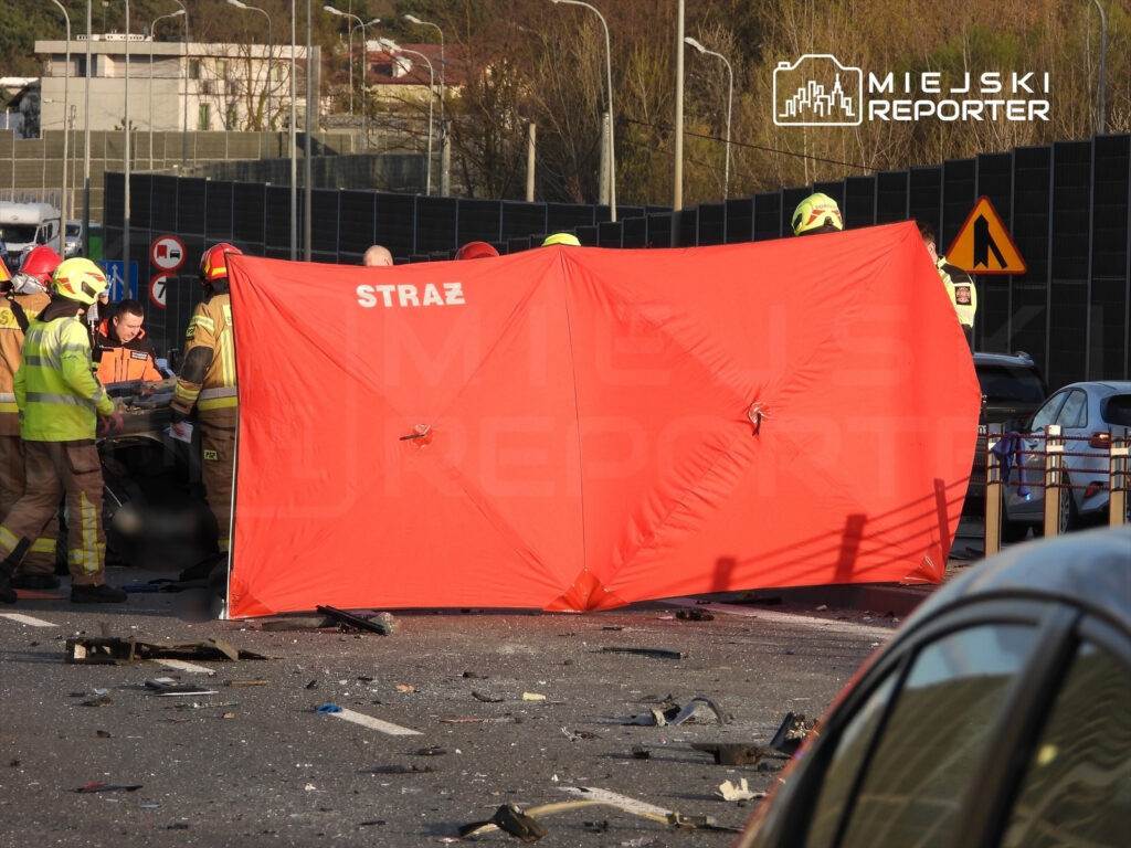 Grupa strażaków pracuje przy wypadku drogowym, zasłaniając miejsce zdarzenia dużym czerwonym namiotem. W tle widać zniszczone pojazdy i rozbite szkło.