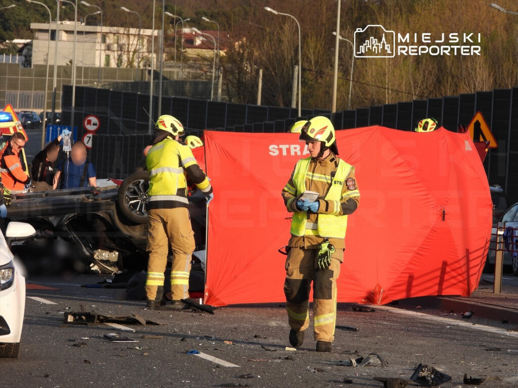 Strażacy zabezpieczają miejsce wypadku drogowego, gdzie przewrócone auto leży na boku, a wokół są rozbite elementy pojazdów.