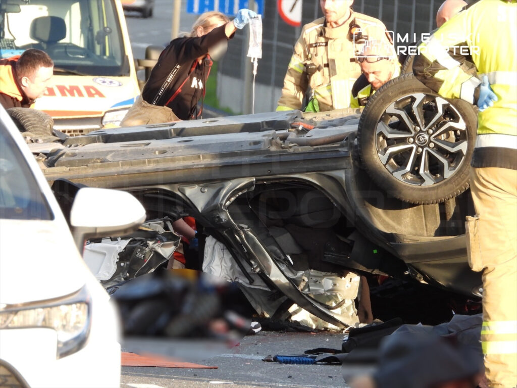 Ratownicy medyczni i strażacy udzielają pomocy poszkodowanym przy przewróconym samochodzie na drodze.