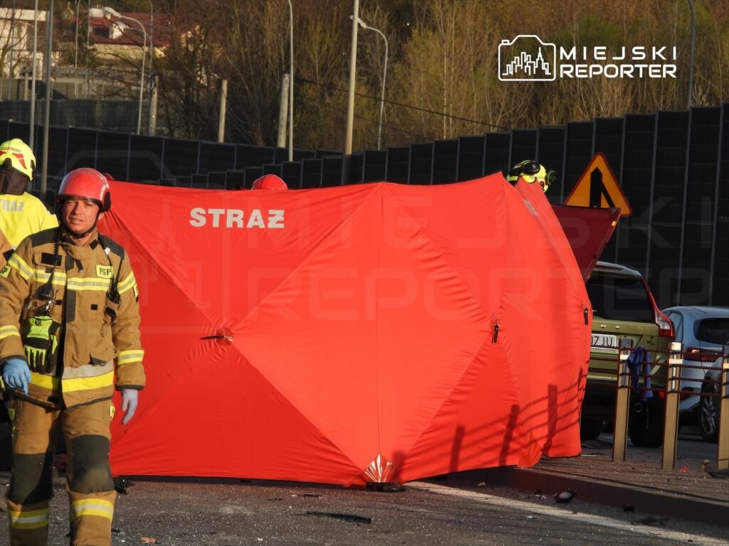 Strażacy w mundurach i hełmach pracują przy wypadku drogowym, zasłaniając miejsce zdarzenia czerwoną osłoną.