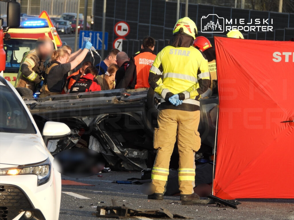 Grupa ratowników medycznych udziela pomocy poszkodowanym przy przewróconym pojeździe na drodze.