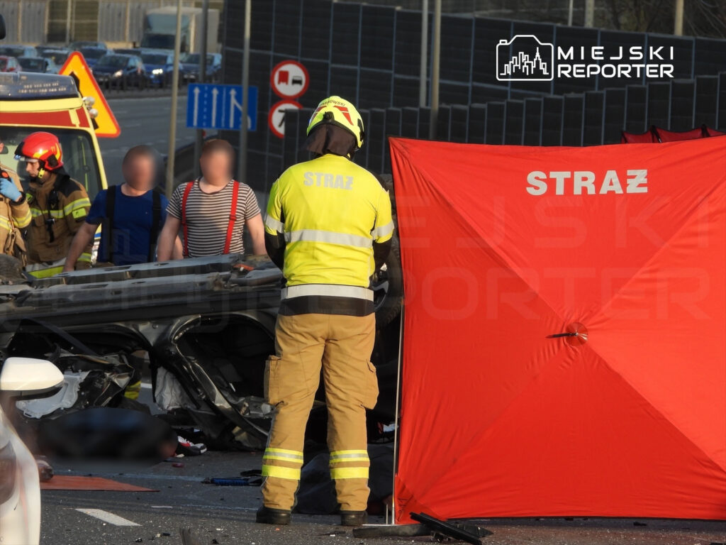 Strażacy w mundurach pracują przy wypadku drogowym, zabezpieczając miejsce zdarzenia czerwoną kotarą.