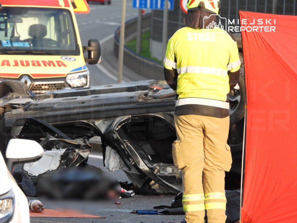 Strażak w żółtej kurtce stoi obok przewróconego samochodu na drodze, a w tle widać karetkę pogotowia.