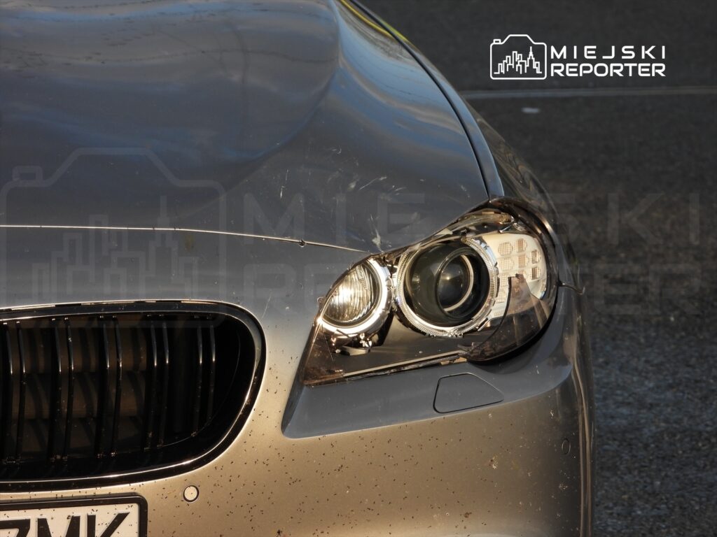 Szary samochód osobowy z uszkodzoną przednią lampą i zanieczyszczoną karoserią, stojący na asfaltowej nawierzchni.