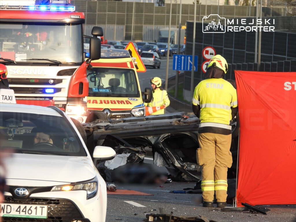 Na drodze służby ratunkowe pracują przy wypadku drogowym, obok przewróconego pojazdu.