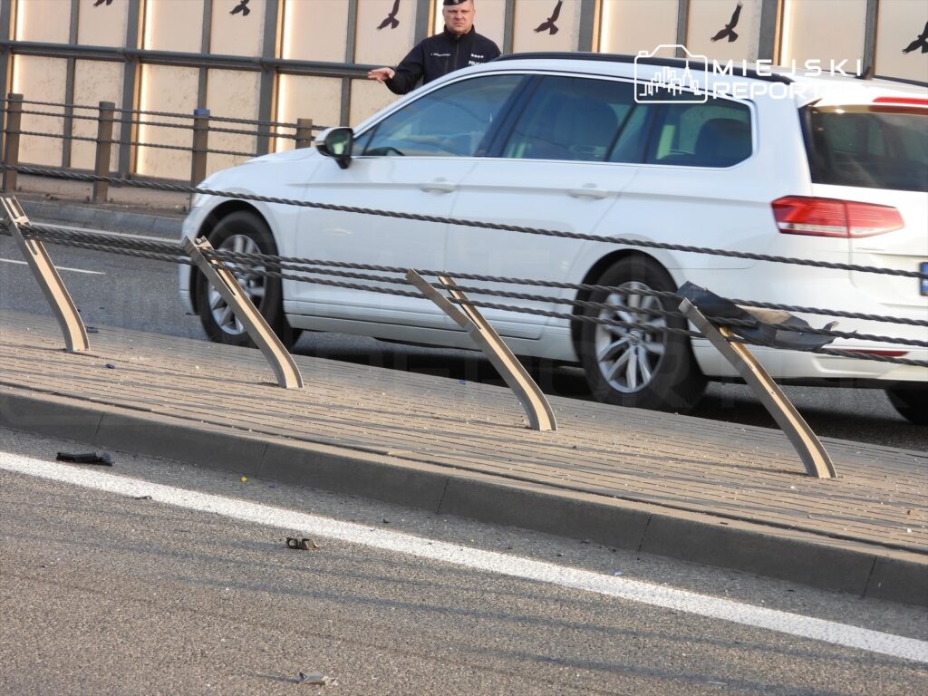 Mężczyzna w granatowej kurtce stoi obok białego samochodu osobowego na ruchliwej ulicy, w tle widoczne barierki.