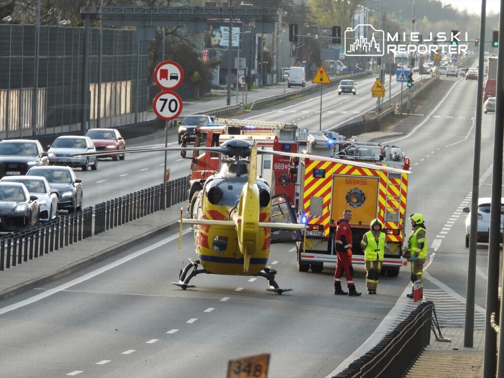Śmigłowiec ratunkowy ląduje na jezdni, obok stoją ratownicy medyczni i strażacy, a ruch drogowy jest utrudniony.