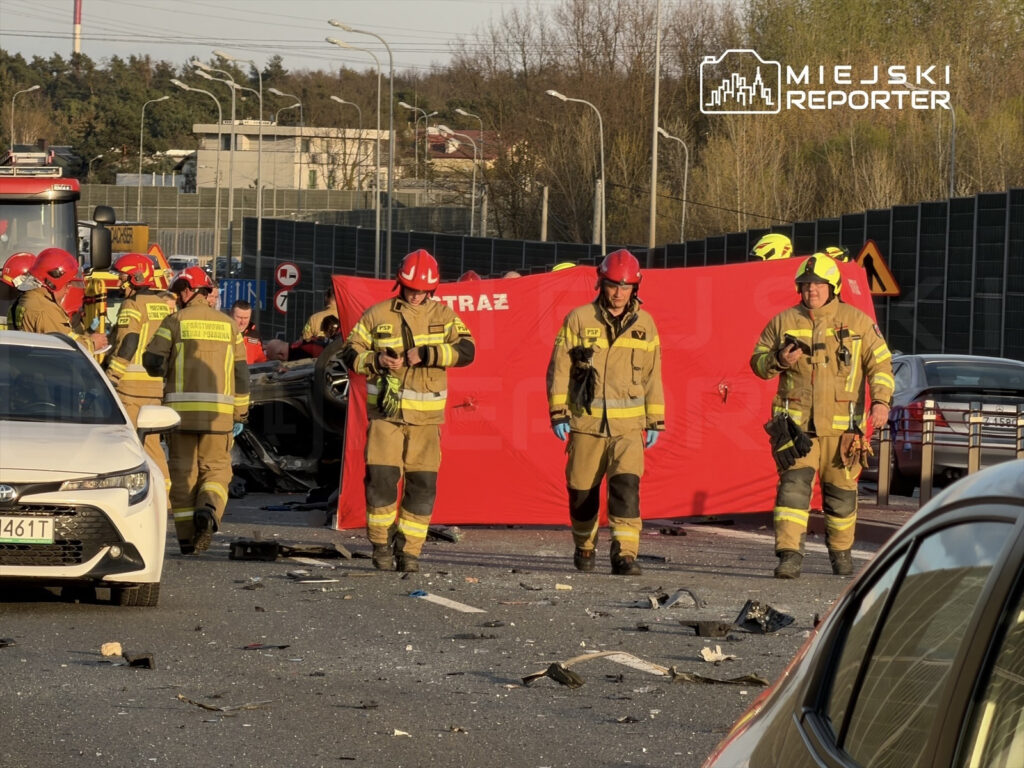 Grupa strażaków w hełmach i mundurach przeszukuje miejsce wypadku drogowego, obok przewróconego samochodu.