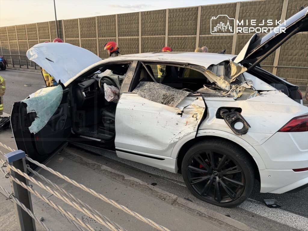 Uszkodzony samochód osobowy z otwartą maską i zniszczonym tyłem, obok pracujący strażacy.