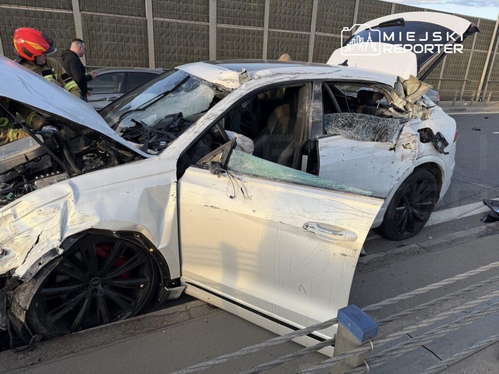 Uszkodzony biały samochód osobowy na drodze, z otwartymi drzwiami i zniszczoną karoserią, obok stoją strażacy.
