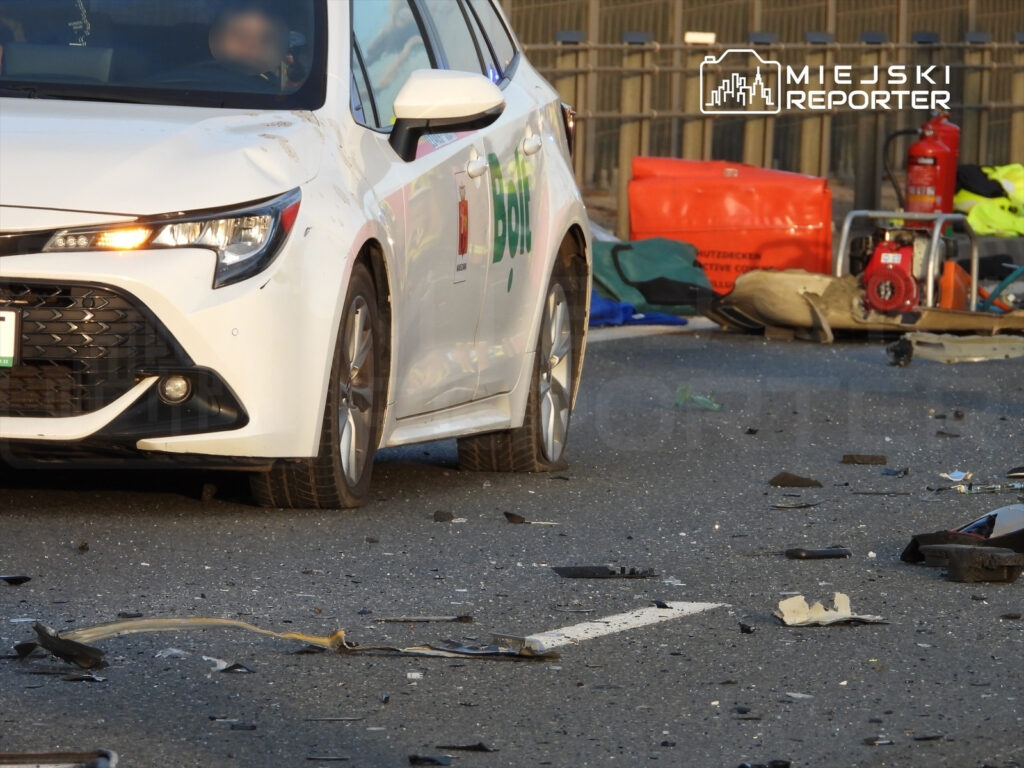 Biały samochód osobowy stoi na drodze, wokół leżą rozbite elementy i odpadki po wypadku.