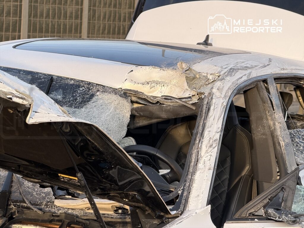 Uszkodzony samochód z wgniecionym dachem i rozbitymi szybami, stojący na parkingu.