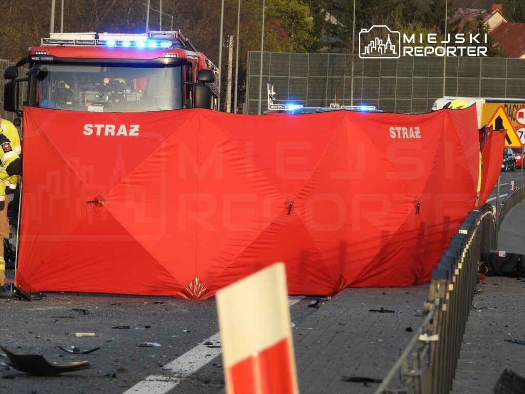 Strażacy ustawiają czerwoną osłonę na drodze w miejscu wypadku, podczas gdy pojazd strażacki z włączonymi sygnałami świetlnymi stoi w tle.