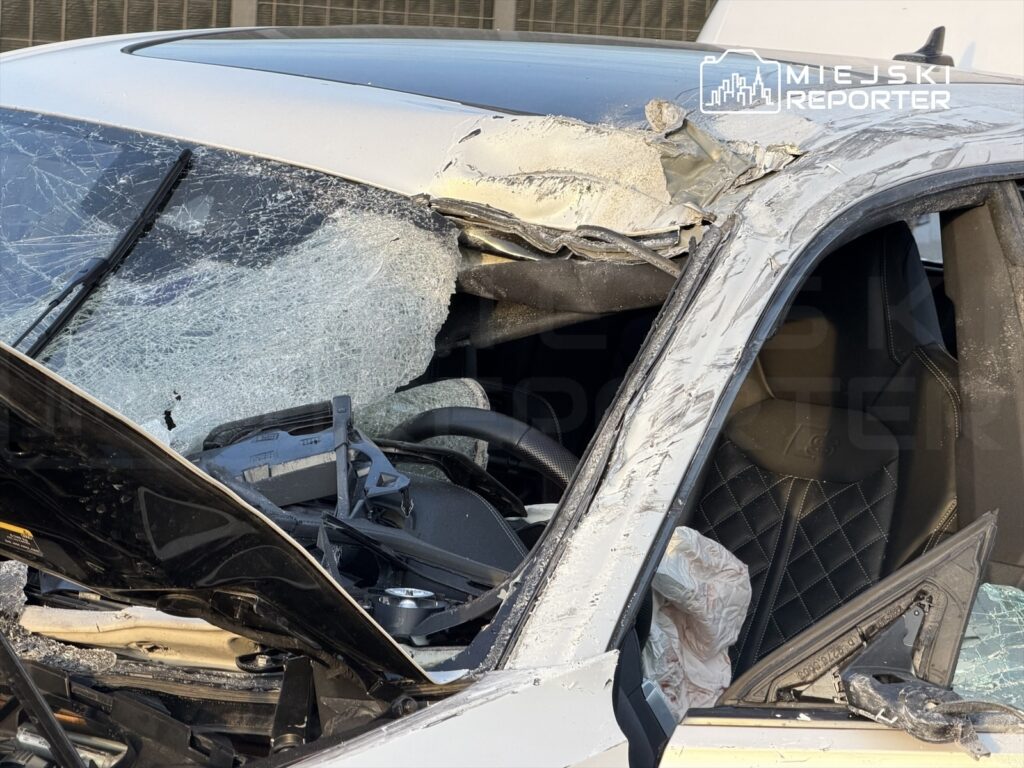 Uszkodzony samochód osobowy z rozbitą szybą przednią i zniekształconym dachem, stojący na parkingu.