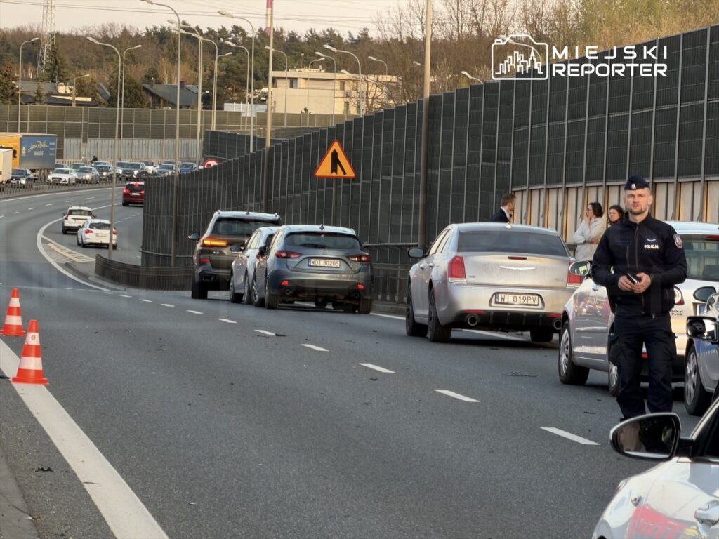Policjant w mundurze stoi na poboczu drogi, a w tle widoczne są zablokowane pojazdy i znak ostrzegawczy.
