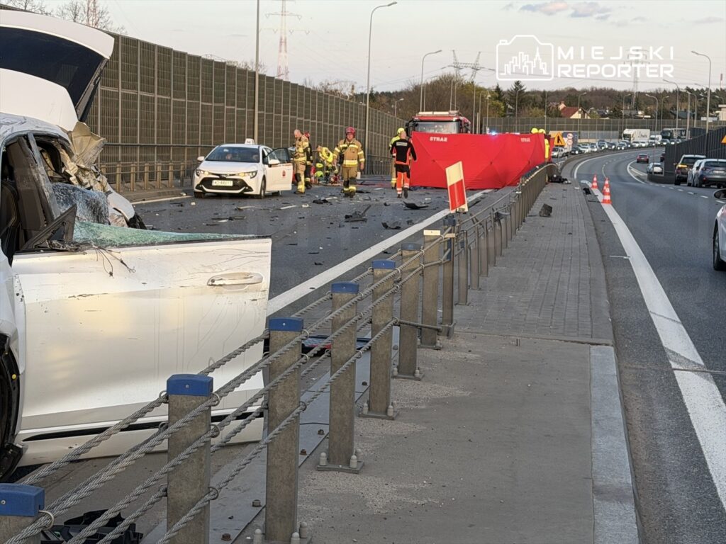 W wyniku wypadku drogowego uszkodzony samochód osobowy leży na jezdni, a obok pracują strażacy i ratownicy medyczni.