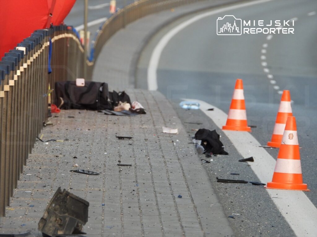 Na jezdni leżą rozbite przedmioty oraz torba, a obok ustawione są pomarańczowe pachołki drogowe.