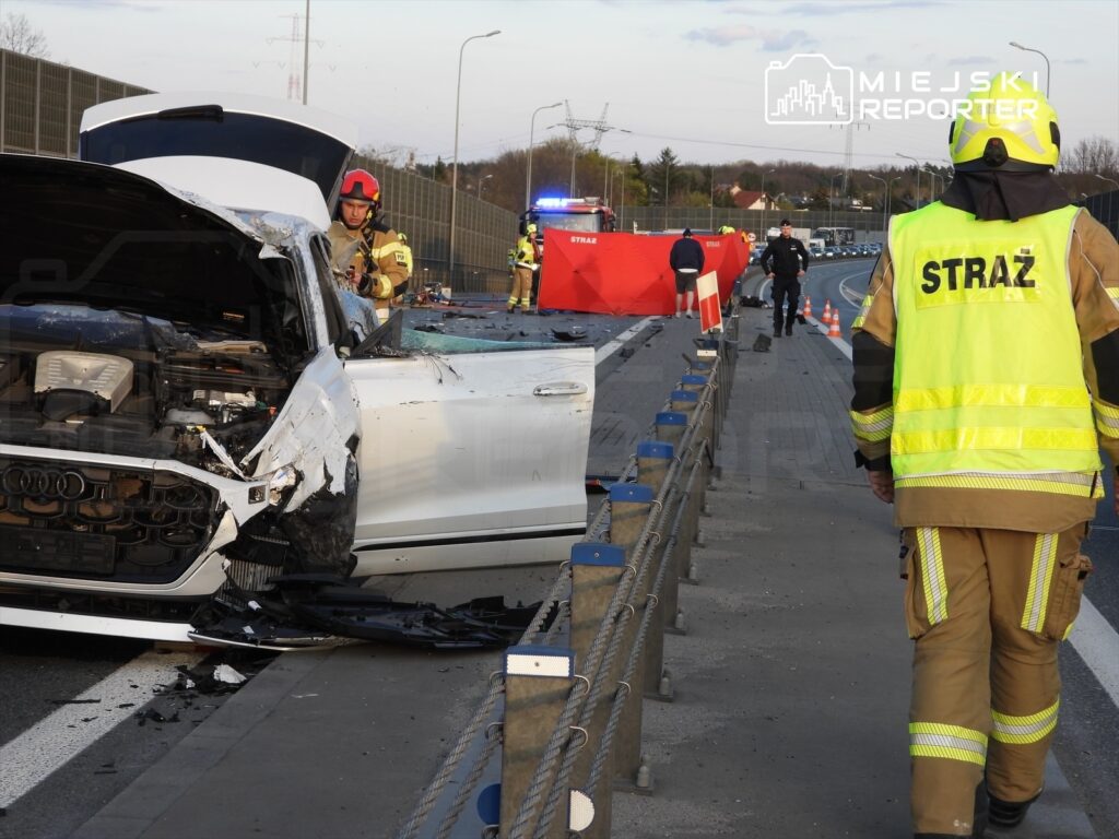 Strażak w żółtej kamizelce podchodzi do uszkodzonego samochodu osobowego na autostradzie, obok widać pojazdy służb ratunkowych.