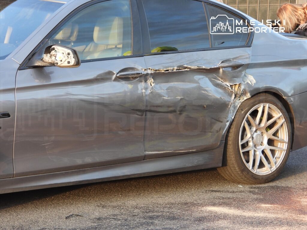 Uszkodzony samochód osobowy z wgniecioną boczną częścią i uszkodzonym lusterkiem zaparkowany na ulicy.