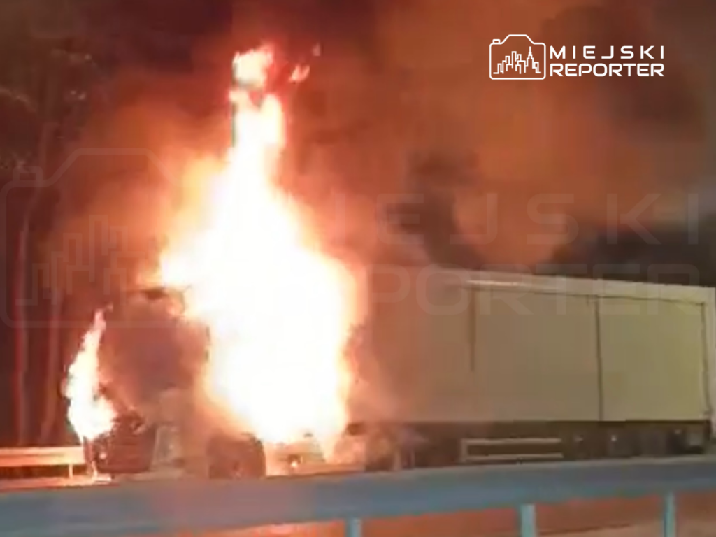 Tir objęty ogniem na poboczu drogi, z intensywnymi płomieniami i dymem unoszącym się w nocy.