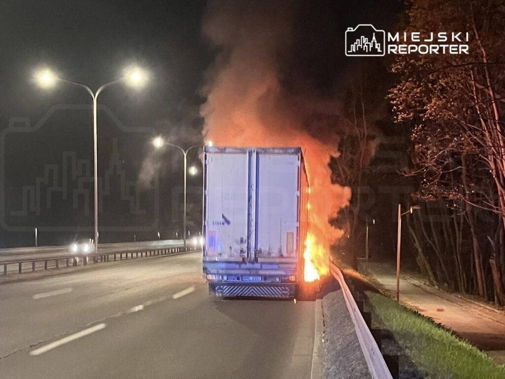 Ciągnik siodłowy zapalił się na poboczu drogi, wydobywając ogień i dym w nocy. W tle widać latarnie uliczne.