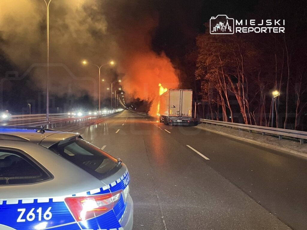 Pojazd ciężarowy płonący na drodze, w tle widoczne płomienie i dym, nocna sceneria.