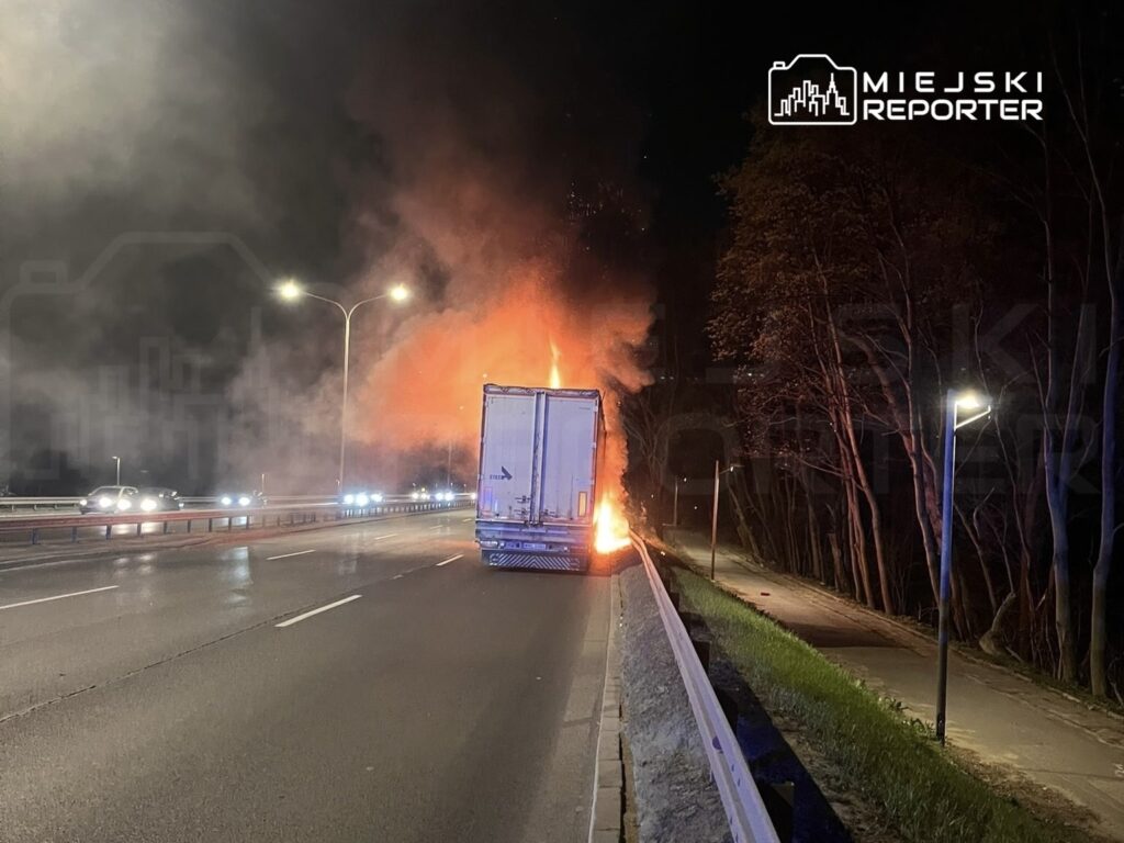 Cysterna płonąca na pasie ruchu, otoczona dymem, przy drodze w nocy. W tle widoczne samochody i drzewa.