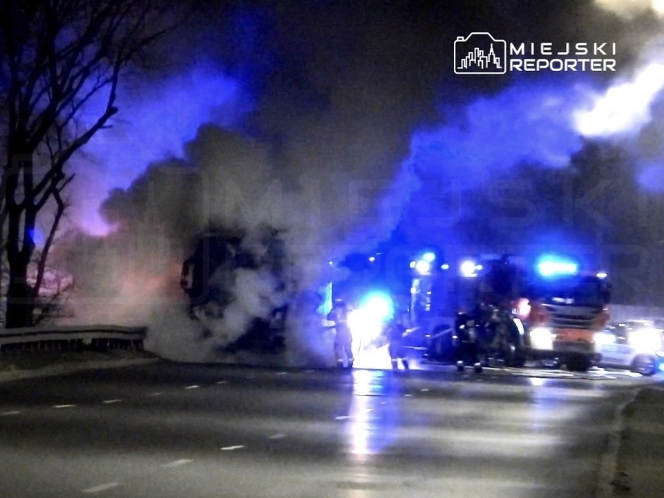 Pożar ciężarówki na drodze, otoczony przez strażaków i pojazdy ratunkowe, w nocy w gęstym dymie.