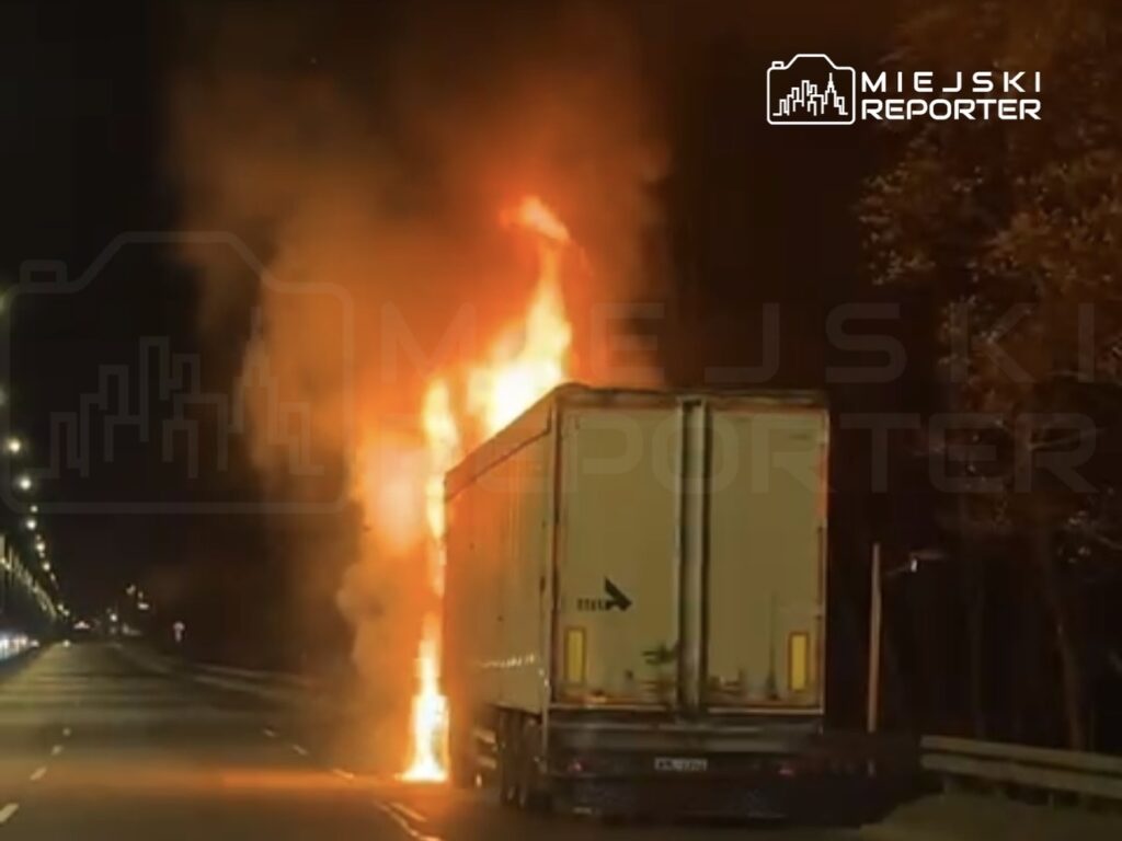 Tir zapalony ogniem na poboczu drogi, w tle nocne oświetlenie ulicy.