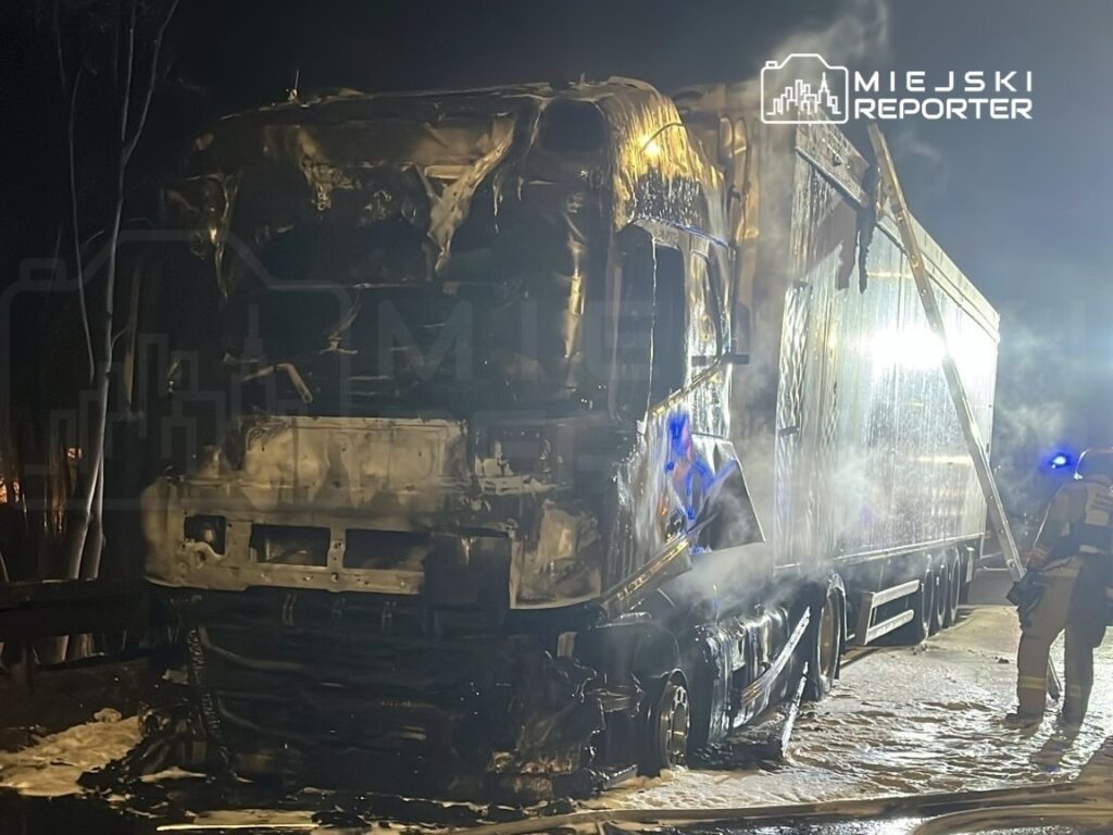 Spalony ciężarówka z uszkodzoną kabiną oraz przyczepą, obok której strażacy gaszą ogień w nocnym otoczeniu.